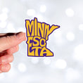 Load image into Gallery viewer, This image shows a hand holding the Minnesota purple and gold sticker.
