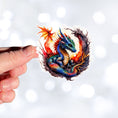 Load image into Gallery viewer, This image shows a hand holding the Baby Dragon in a Nest Die-Cut Sticker.
