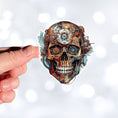 Load image into Gallery viewer, This image shows a hand holding the Steampunk Skull 6 Die-Cut Sticker.
