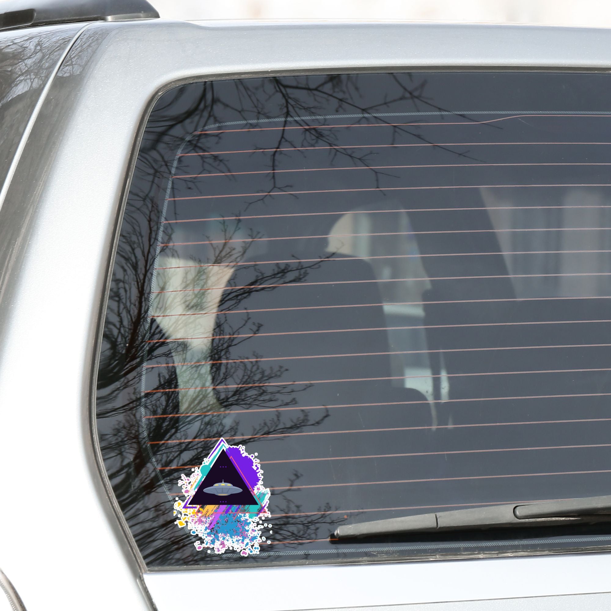 UFO alert! This individual die-cut sticker features an alien spaceship, UFO, on a black triangle background with pastel paint splatters behind. This image shows the alien triangle sticker on the rear window of a car.