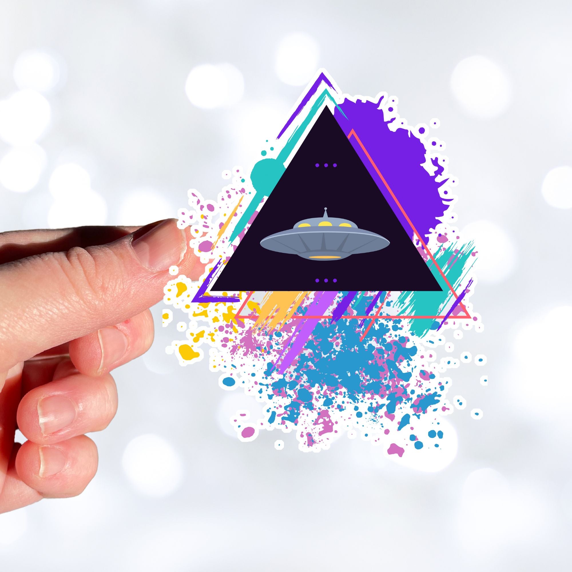 UFO alert! This individual die-cut sticker features an alien spaceship, UFO, on a black triangle background with pastel paint splatters behind. This image shows a hand holding the alien triangle sticker.