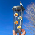 Load image into Gallery viewer, Our Around the World sticker sheet collection has sticker images of iconic travel destinations! This sheet has a red background with 18 different stickers. This image shows a water bottle with stickers of the Golden Gate Bridge, a lighthouse, a suspension bridge, and the Sidney Opera House applied to it.
