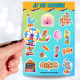 Load image into Gallery viewer, Who doesn't love going to the carnival? This sheet has stickers showing the big top, clowns, animals, and of course your favorite foods like hotdogs and cotton candy. This is an image of a hand holding one of the stickers above the sticker sheet.
