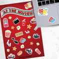 Load image into Gallery viewer, Get your tickets, buy your snacks, and enjoy a night at the movies! This sticker sheet is filled with sticker images like: Popcorn, movie reel, camera, hotdog, soda, and 3D glasses. This image shows the sticker sheet next to an open laptop with stickers of comedy/tragedy masks, and cinema tickets applied below the keyboard. 
