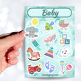 Load image into Gallery viewer, This sticker sheet has stickers showing everything a new parent would want for decorating or scrap booking! Images include a stroller, baby bottle, rocking horse, and even a baby elephant! This image shows a hand holding one of the stickers above the sticker sheet
