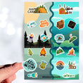 Load image into Gallery viewer, Get away to the woods, mountains, or beach with this sticker sheet! This sticker sheet has sticker images for camping in a tent or camper, or in a cabin or cottage. This image shows a hand holding a sticker reading "Camping Crew" above the sticker sheet.
