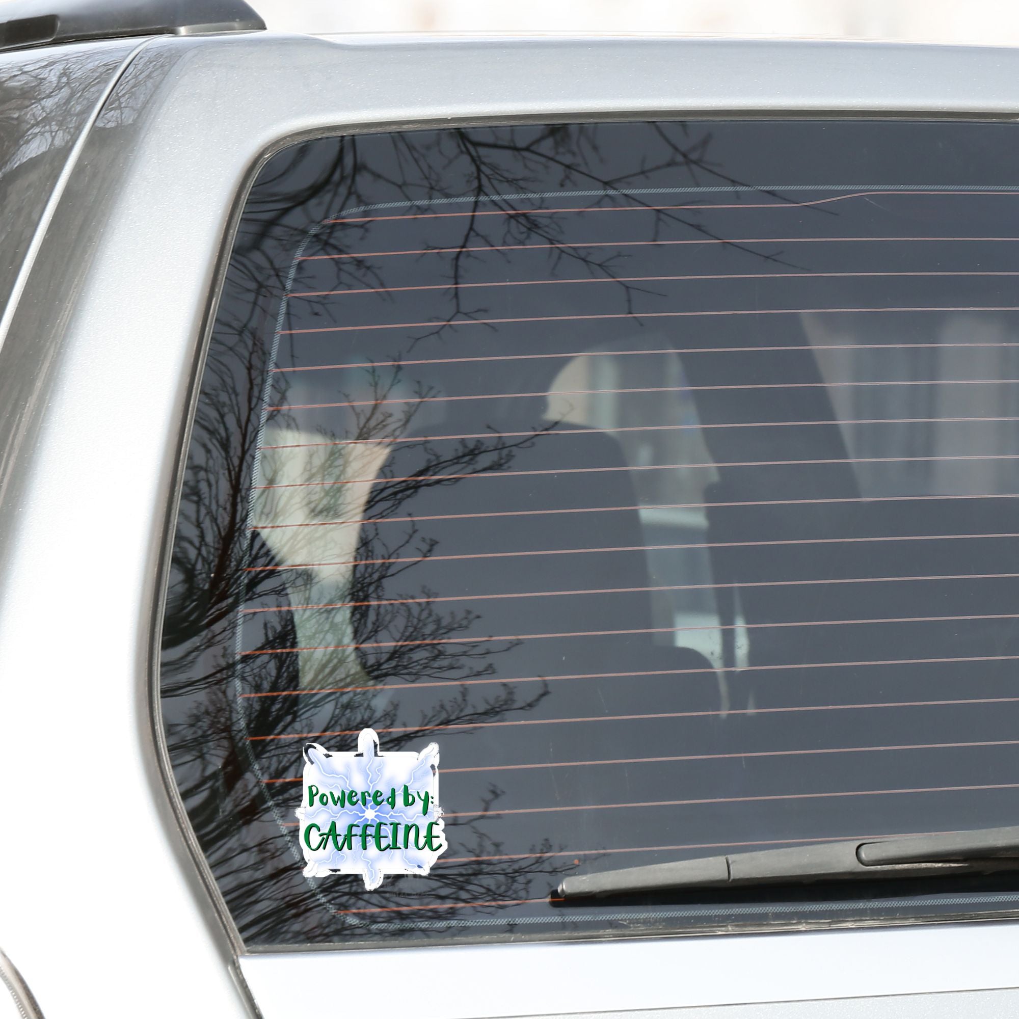 For those who like it, caffeine can be the magic elixir! This individual die-cut sticker has the words Powered by CAFFEINE in green over a background of blue electric/lightning bolts. This image shows the Powered by Caffeine sticker on the back window of a car. 