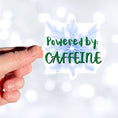Load image into Gallery viewer, For those who like it, caffeine can be the magic elixir! This individual die-cut sticker has the words Powered by CAFFEINE in green over a background of blue electric/lightning bolts. This image shows a hand holding the Powered by Caffeine sticker.
