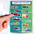 Load image into Gallery viewer, This sticker sheet has a collection of car and truck stickers; perfect for kids and adults who like their four wheel friends! This sheet has six different car stickers and six different truck stickers on a green and blue background. This image shows a hand holding a race car sticker over the sticker sheet.
