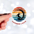 Load image into Gallery viewer, Hang 10 Bro! Catch a wave with this individual die-cut sticker featuring the silhouette of a male surfer inside the tube, all on a colored gradient background. Cowabunga! This image shows a hand holding the Catching a Wave sticker. 
