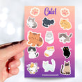 Load image into Gallery viewer, This sticker sheet is purrfect for all cat lovers! It has stickers of 12 different cute and cuddly cats, plus two pawprint stickers.  This image shows a hand holding a brown cat sticker above the full sticker sheet.
