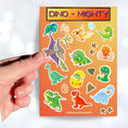 Load image into Gallery viewer, These dinosaur stickers are a-roar-able! This sticker sheet has all of your favorite dinosaurs plus dino eggs, plants, and dino footprints. This image shows a hand holding a tetradactyl sticker over the sticker sheet. 
