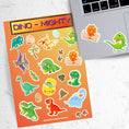 Load image into Gallery viewer, These dinosaur stickers are a-roar-able! This sticker sheet has all of your favorite dinosaurs plus dino eggs, plants, and dino footprints. This image has the sticker sheet next to an open laptop with a sticker of a dino hatching from and egg and a T-Rex dino sticker applied below the keyboard.
