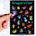 Load image into Gallery viewer, This sticker sheet is filled with cute images of baby dragons, and of course their treasure! This image shows a hand holding a sticker of a purple dragon sitting with butterflies flying around over the sticker sheet.
