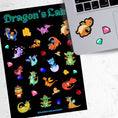 Load image into Gallery viewer, This sticker sheet is filled with cute images of baby dragons, and of course their treasure! This image shows the sticker sheet next to an open laptop with a sticker of an orange dragon with green wings and two gem stickers applied below the keyboard.
