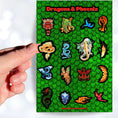 Load image into Gallery viewer, Set on a background of green scales, this sticker sheet has 16 different stickers of dragons and phoenix. This image shows a hand holding a feathery golden dragon above the sticker sheet.
