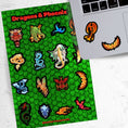 Load image into Gallery viewer, Set on a background of green scales, this sticker sheet has 16 different stickers of dragons and phoenix. This image shows the sticker sheet next to an open laptop with two phoenix stickers applied below the keyboard.
