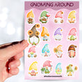 Load image into Gallery viewer, Gnomes, gnomes, and more gnomes! This sticker sheet has 16 unique gnome stickers for people who love their Gnomies. This image shows a hand holding a sticker of a gnome with sunflowers in her hair above the sticker sheet.
