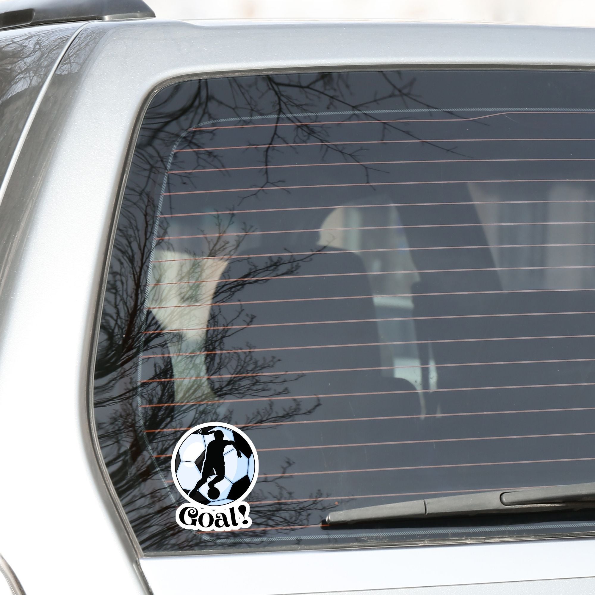 Show your love of soccer, or football, with this individual die-cut sticker! This sticker shows the silhouette of a player with a ponytail about to kick, on a black and white/blue soccer ball background, with the word "Goal!" below. This image shows the soccer sticker on the rear window of a car.