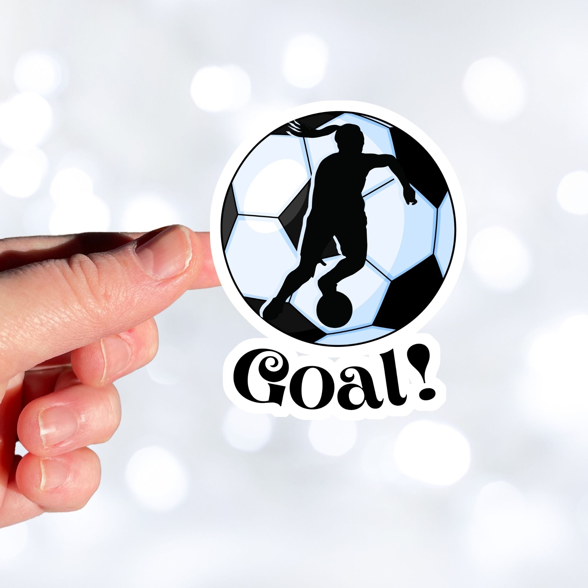 Show your love of soccer, or football, with this individual die-cut sticker! This sticker shows the silhouette of a player with a ponytail about to kick, on a black and white/blue soccer ball background, with the word "Goal!" below. This image shows a hand holding the soccer sticker.
