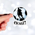 Load image into Gallery viewer, Show your love of soccer, or football, with this individual die-cut sticker! This sticker shows the silhouette of a player about to kick, on a black and white/blue soccer ball background, with the word "Goal!" below. This image shows a hand holding the soccer sticker.
