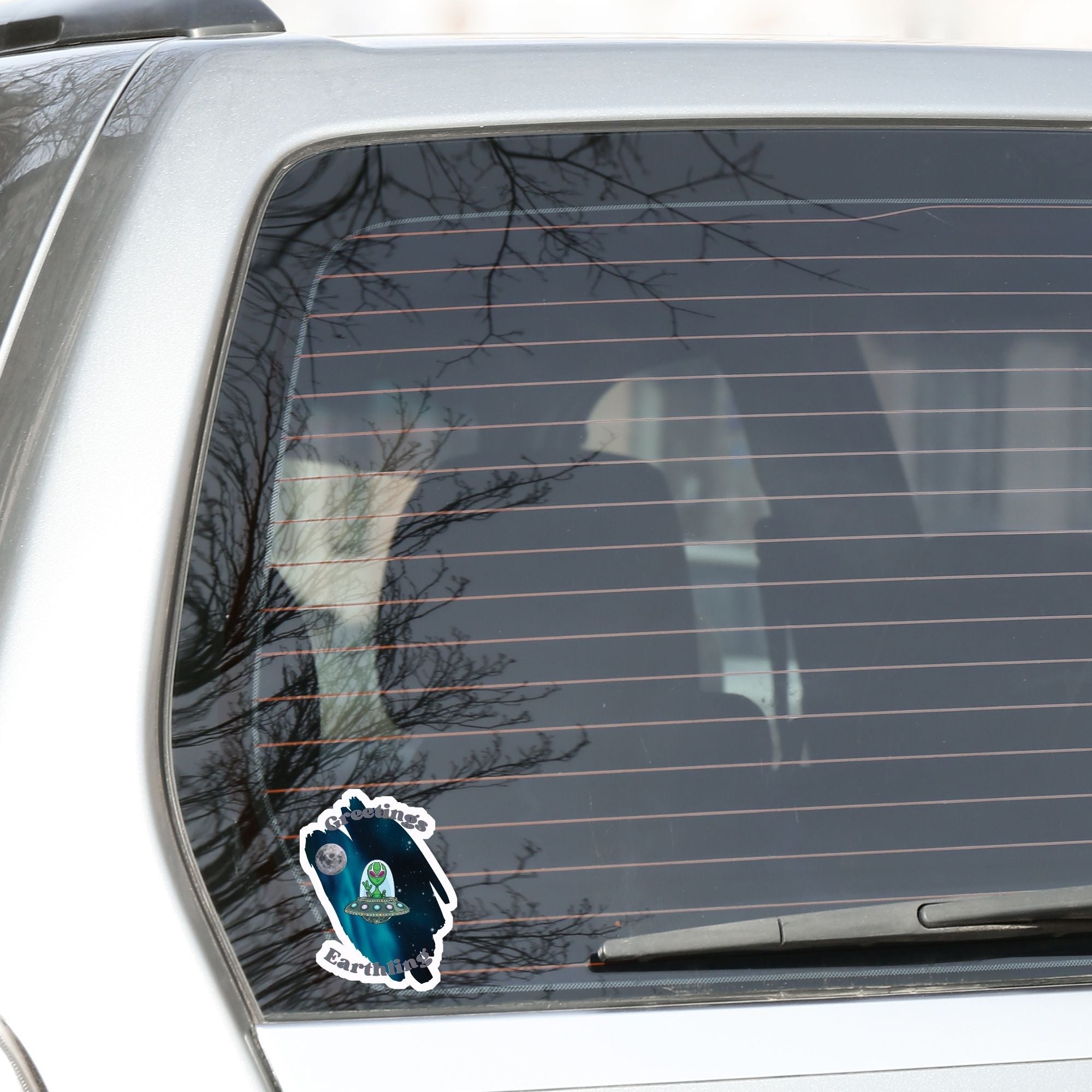 Welcome space aliens! This individual die-cut sticker features a friendly green space alien in a flying saucer, on a background of stars with the moon behind, and the words "Greetings Earthling". This image shows the flying saucer sticker on the rear window of a car.