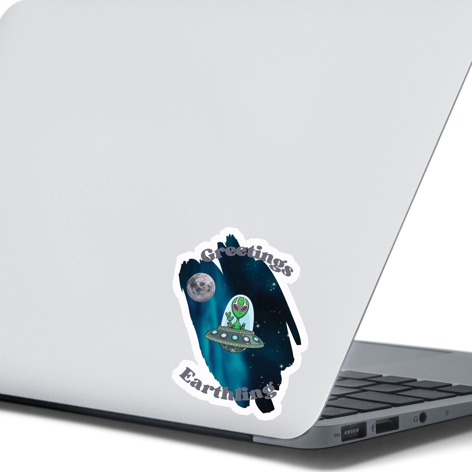 Welcome space aliens! This individual die-cut sticker features a friendly green space alien in a flying saucer, on a background of stars with the moon behind, and the words "Greetings Earthling". This image shows the flying saucer sticker on the back of an open laptop.