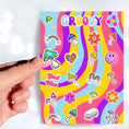 Load image into Gallery viewer, Groovy man! This psychedelic sticker sheet is filled with images of rainbows, mushrooms, flowers, a smiley face, and even a VW Microbus! This image shows a hand holding a blue and green topped mushroom over the sticker sheet.
