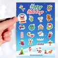 Load image into Gallery viewer, Celebrate the holidays with this winter themed sticker sheet. With sticker images of Santa, elves, snowmen, and your favorite holiday treats, this sticker sheet will bring holiday cheer to every household! It also features a holo star overlay for that extra sparkle! This image shows a hand holding a sticker of Santa snow globe over the sticker sheet. 
