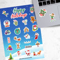 Load image into Gallery viewer, Celebrate the holidays with this winter themed sticker sheet. With sticker images of Santa, elves, snowmen, and your favorite holiday treats, this sticker sheet will bring holiday cheer to every household! It also features a holo star overlay for that extra sparkle! This image shows the sticker sheet next to an open laptop with stickers of a holiday owl and an elf applied below the keyboard.
