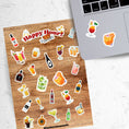 Load image into Gallery viewer, Cheers! Filled with sticker images of your favorite cocktails and adult beverages, this is for adults who can enjoy this sticker sheet responsibly! This image shows the sticker sheet next to an open laptop with stickers of mixed drinks applied below the keyboard.
