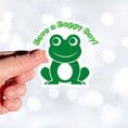 Load image into Gallery viewer, This inspirational sticker features a smiling frog with "Have a Hoppy Day!" written above. This image shows a hand holding the Have a Hoppy Day sticker.
