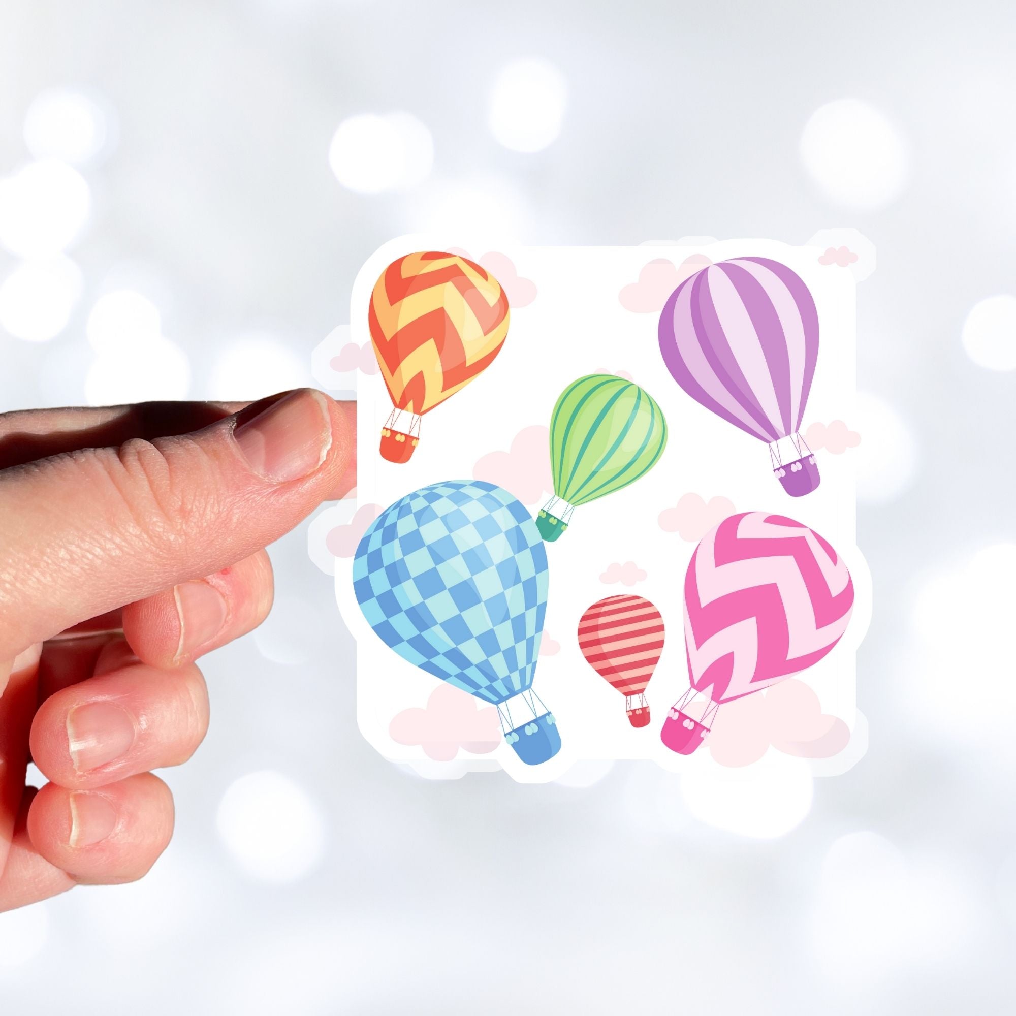 Float away on a colorful hot air balloon! This individual die-cut hot air balloon sticker has six, yes six, different hot air balloons on a background of clouds. This image shows a hand holding the hot air balloon sticker.