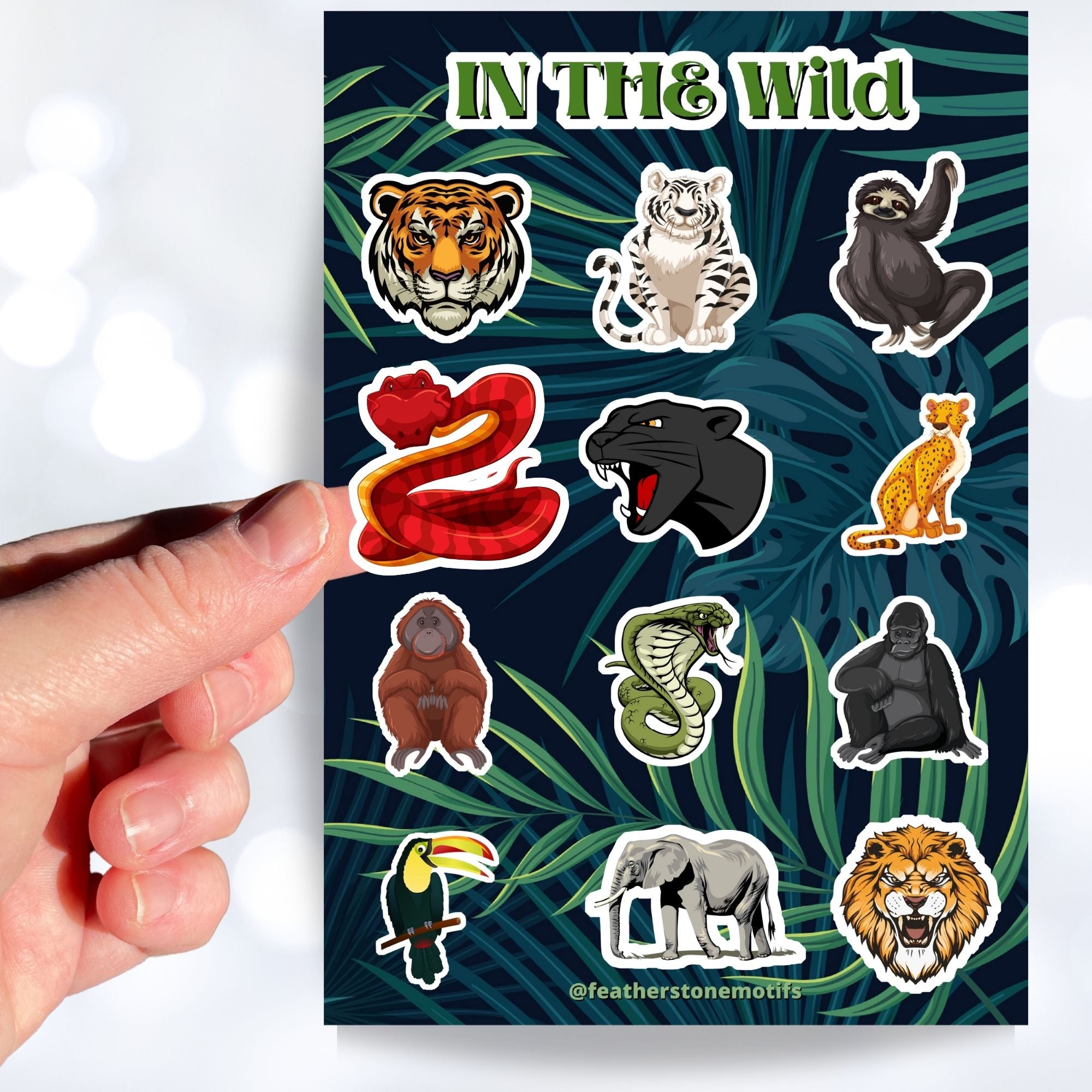 This sticker sheet has sticker images of your favorite jungle and rain forest creatures! There are a dozen different sticker images including tigers, a lion, a gorilla, snakes, and even a sloth.  This image shows a hand holding a sticker of a snake over the sticker sheet.