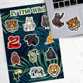 Load image into Gallery viewer, This sticker sheet has sticker images of your favorite jungle and rain forest creatures! There are a dozen different sticker images including tigers, a lion, a gorilla, snakes, and even a sloth.  This image shows the sticker sheet next to an open laptop with stickers of a sloth and a tiger head applied below the keyboard.
