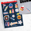 Load image into Gallery viewer, Celebrate summer, and the 4th of July in the USA. This sticker sheet has all of your favorite summer time foods like BBQing, burgers, hot dogs, and corn on the cob. Plus fireworks and red, white, and blue images. This image has the sticker sheet next to an open laptop with USA and a summer grill stickers.
