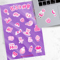 Load image into Gallery viewer, Congratulations, it's a girl! This sticker sheet is perfect for parents welcoming a baby girl into their family and it has lots of images of toys, a bottle, a stroller, and animals, all in pink on a purple background. This image shows the sticker sheet next to an open laptop with stickers of a stroller with an elephant and balloons, a moon and stars crib mobile, and a heart with BABY written on it, applied below the keyboard.
