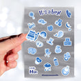 Load image into Gallery viewer, Congratulations, it's a boy! This sticker sheet is perfect for parents welcoming a baby boy into their family and it has lots of images of toys, a bottle, a stroller, and animals, all in blue on a gray background. This image shows a hand holding a sticker of a xylophone above the sticker sheet. 
