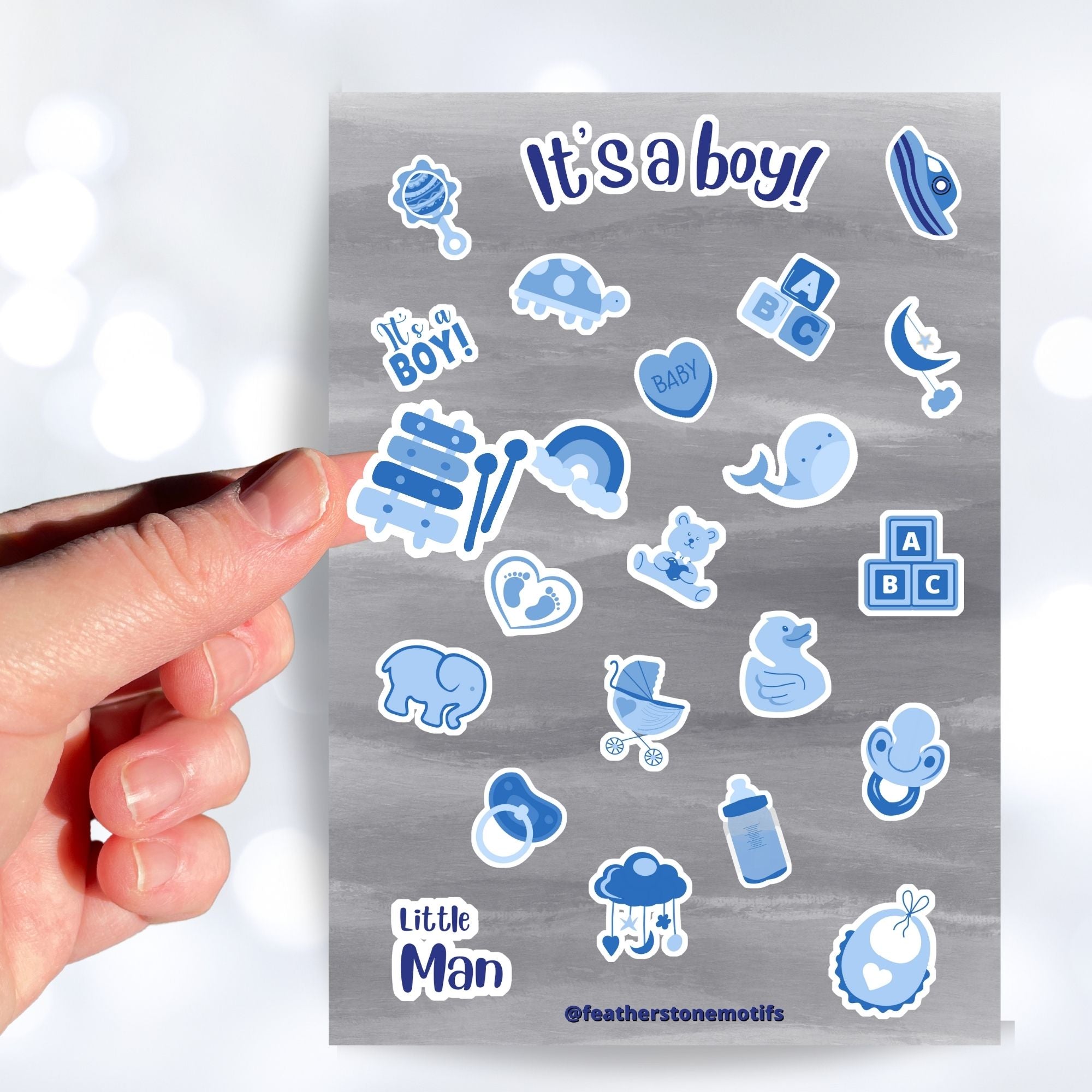 Congratulations, it's a boy! This sticker sheet is perfect for parents welcoming a baby boy into their family and it has lots of images of toys, a bottle, a stroller, and animals, all in blue on a gray background. This image shows a hand holding a sticker of a xylophone above the sticker sheet. 