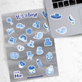 Load image into Gallery viewer, Congratulations, it's a boy! This sticker sheet is perfect for parents welcoming a baby boy into their family and it has lots of images of toys, a bottle, a stroller, and animals, all in blue on a gray background. This image shows the sticker sheet next to an open laptop with stickers of a boat, a moon and stars crib mobile, and a whale, applied below the keyboard.
