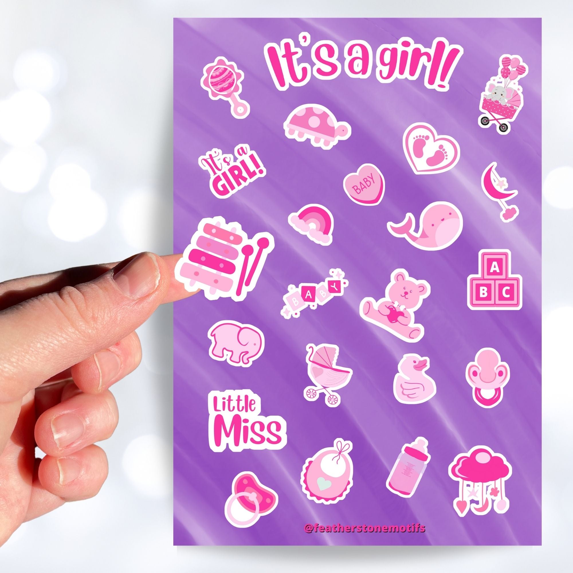 Congratulations, it's a girl! This sticker sheet is perfect for parents welcoming a baby girl into their family and it has lots of images of toys, a bottle, a stroller, and animals, all in pink on a purple background. This image shows a hand holding a xylophone sticker above the sticker sheet.
