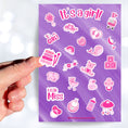 Load image into Gallery viewer, Congratulations, it's a girl! This sticker sheet is perfect for parents welcoming a baby girl into their family and it has lots of images of toys, a bottle, a stroller, and animals, all in pink on a purple background. This image shows a hand holding a xylophone sticker above the sticker sheet.
