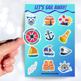 Load image into Gallery viewer, Permission to come aboard and sail away? This sticker sheet has twelve different sailing designs perfect for any captain or first mate! This image shows a hand holding a compass sticker above the sticker sheet.
