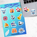 Load image into Gallery viewer, Permission to come aboard and sail away? This sticker sheet has twelve different sailing designs perfect for any captain or first mate! This image shows the sticker sheet next to an open laptop with a nautical owl sticker and a sticker of a life ring, telescope, and ships wheel applied below the keyboard.
