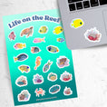 Load image into Gallery viewer, Teaming with sea life, this sticker sheet is filled with sticker images of fish, sea turtles, and coral! Take the reef with you anywhere with these beautiful stickers. This image shows the sticker sheet next to an open laptop with stickers of a yellow angle fish and coral reef applied below the keyboard.
