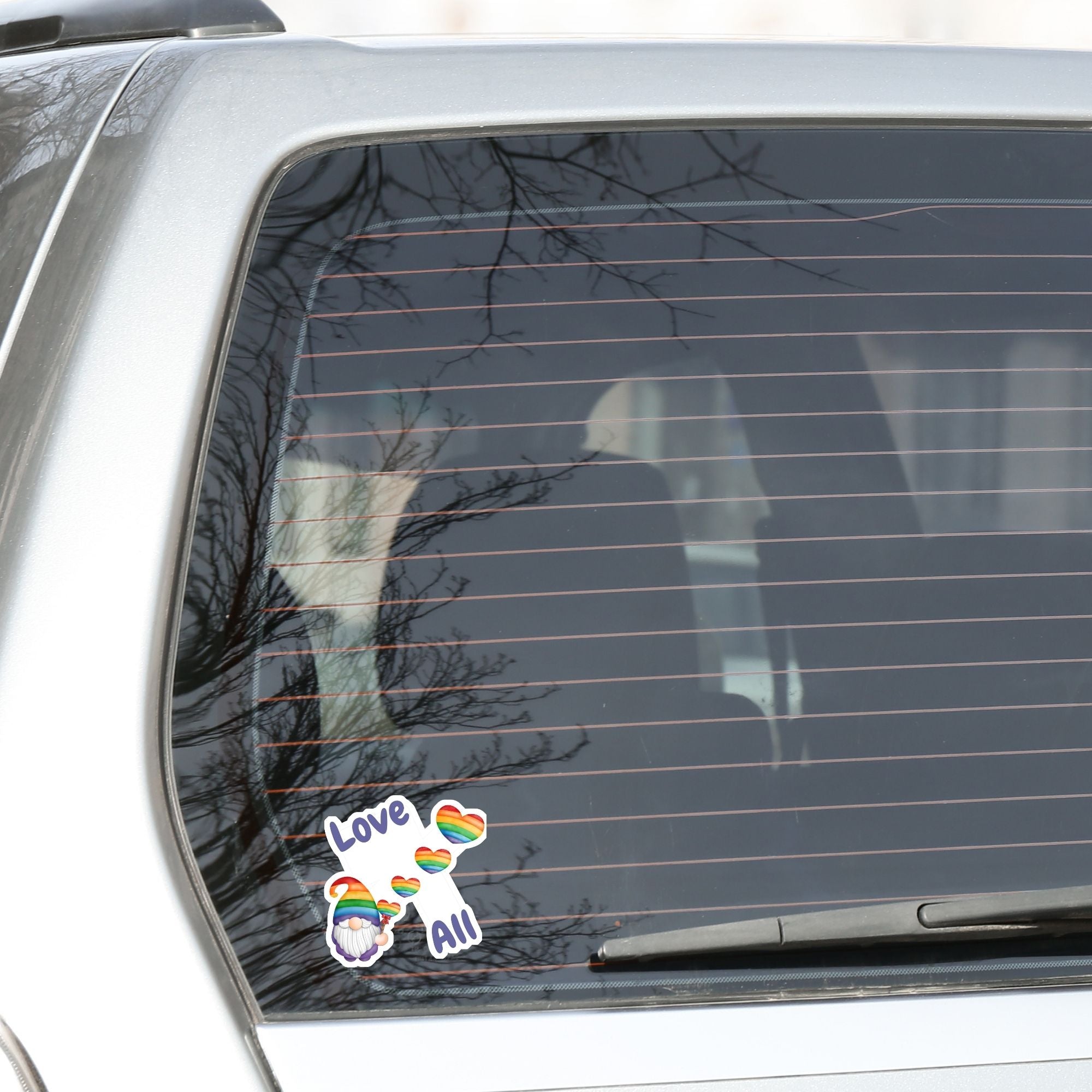 This gnome is showing his Pride! With the words "Love All", this individual die-cut sticker features a gnome with a rainbow hat and rainbow heart balloons. This image shows the Gnome Love All die-cut sticker on the back window of a car.