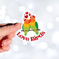Load image into Gallery viewer, Aww, Love Birds! This individual die-cut sticker features a pair of love birds with hearts above. This is a perfect gift for your love bird! This image shows a hand holding the Love Birds sticker.
