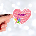 Load image into Gallery viewer, Show how much you love your mom with this individual die-cut sticker. This makes a great gift for mom, or for all mom's to proudly show they are a mother! This sticker features a pink scribbled heart with Mom written across the middle and 5 small paint hands on the lower left side. This image shows a hand holding the Mom sticker.
