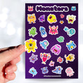 Load image into Gallery viewer, Don't look under the bed! No - these monster's aren't scary! This sticker sheet is filled with sticker images of cute and fun monsters that kids aged 1 to 100 will enjoy. This image shows a hand holding a yellow one eyed monster sticker above the sticker sheet.
