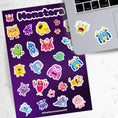 Load image into Gallery viewer, Don't look under the bed! No - these monster's aren't scary! This sticker sheet is filled with sticker images of cute and fun monsters that kids aged 1 to 100 will enjoy. This image shows the sticker sheet next to an open laptop with a sticker of a three eyed green and purple monster sticker and a sad looking blue monster with yellow horns sticker applied below the keyboard.
